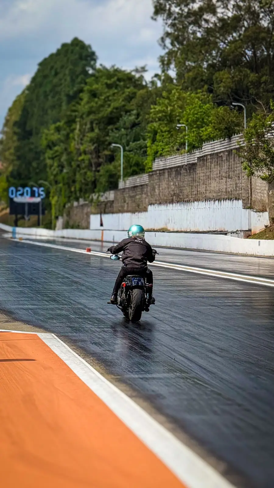 Piloto sozinho acelerando pela pista molhada, visto de trás, com a faixa laranja da borda em primeiro plano.