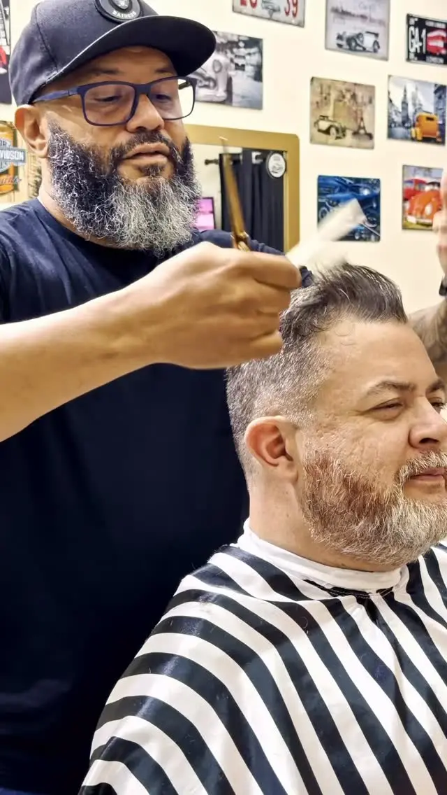 Barbeiro finalizando o corte de um cliente na cadeira, com a parede de quadros da barbearia ao fundo.