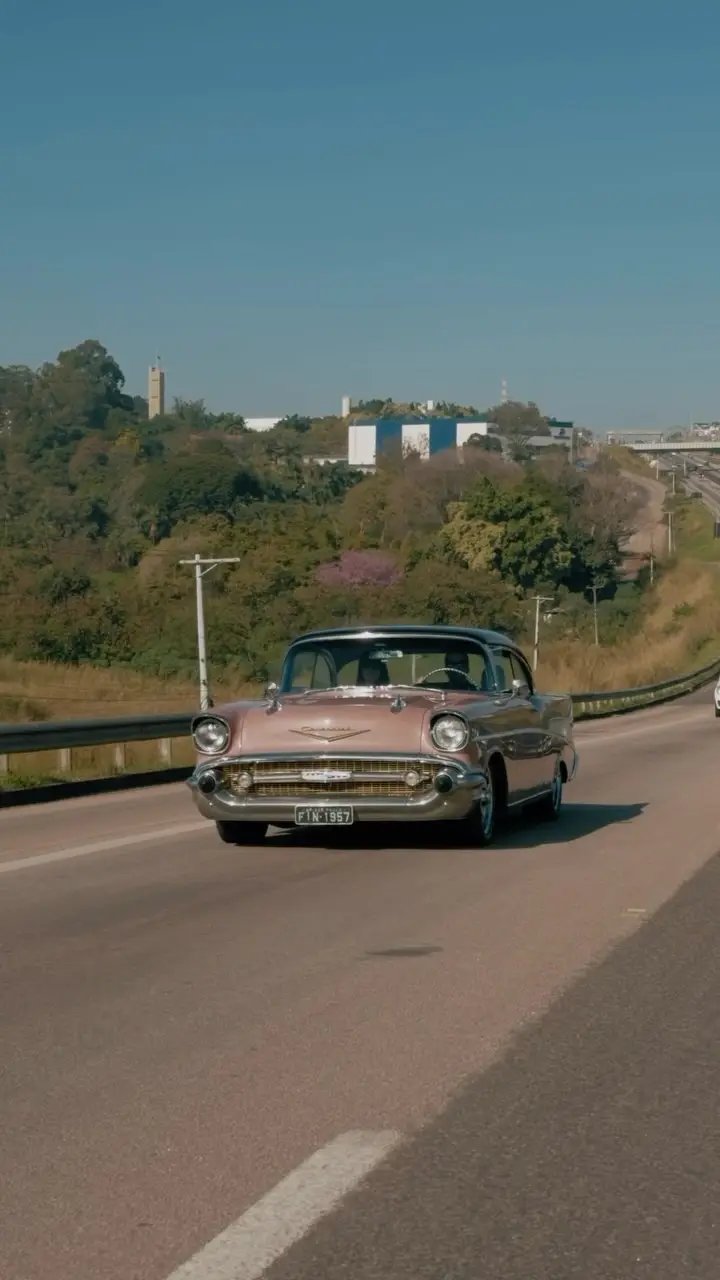 Carro antigo cor-de-rosa descendo uma rodovia arborizada em dia de sol.