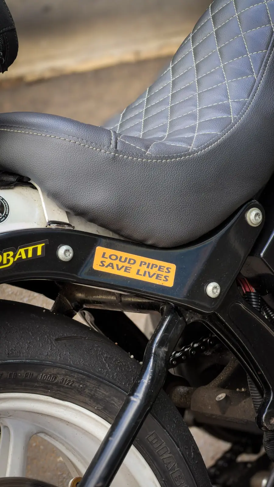 Detalhe do banco costurado de uma Harley-Davidson, com um adesivo Loud Pipes Save Lives no chassi.