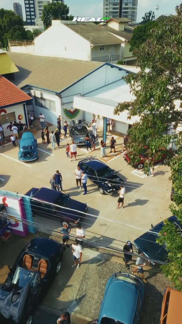 Vista aérea de um encontro de carros, com veículos estacionados em fila e público circulando entre eles.