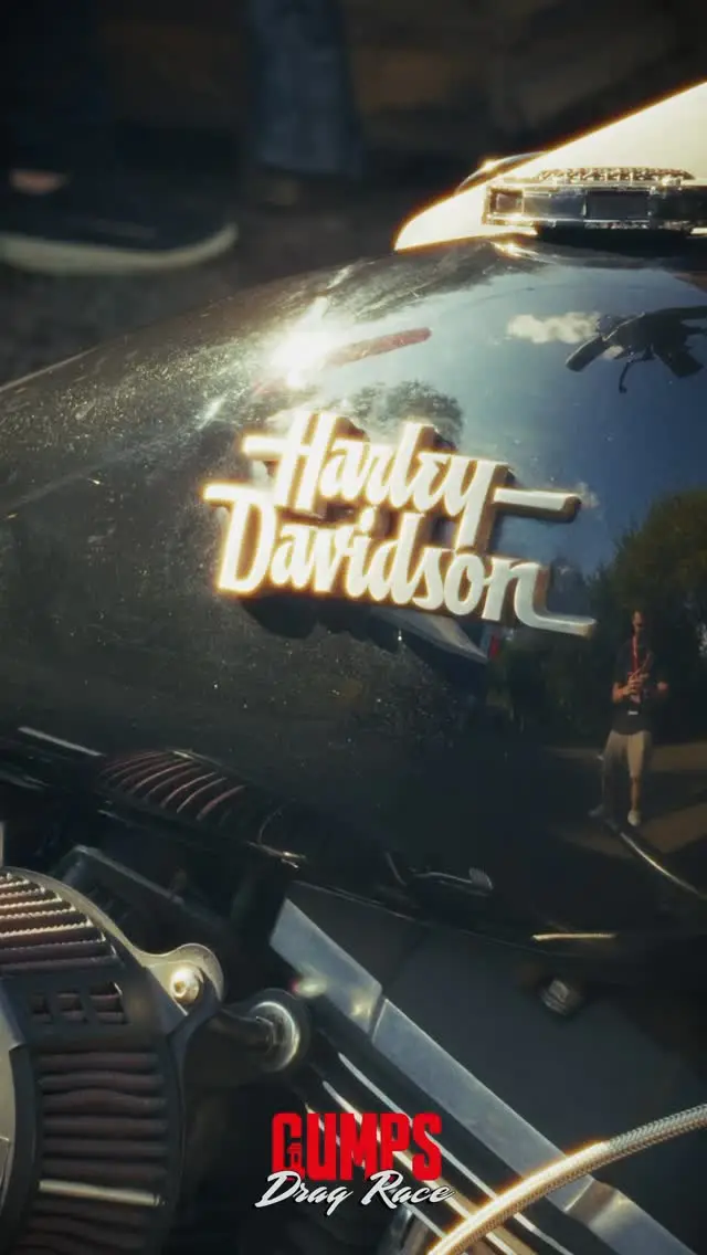 Detalhe do tanque preto de uma Harley-Davidson refletindo a luz, com o logotipo do Gumps Drag Race no canto.