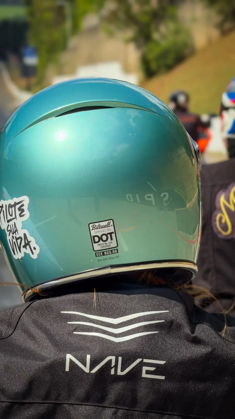 Costas da jaqueta preta de um piloto de capacete verde, parado de frente para a pista.