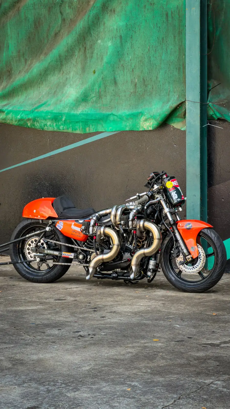 Moto de arrancada laranja parada no box, com os quatro escapamentos cromados à mostra.
