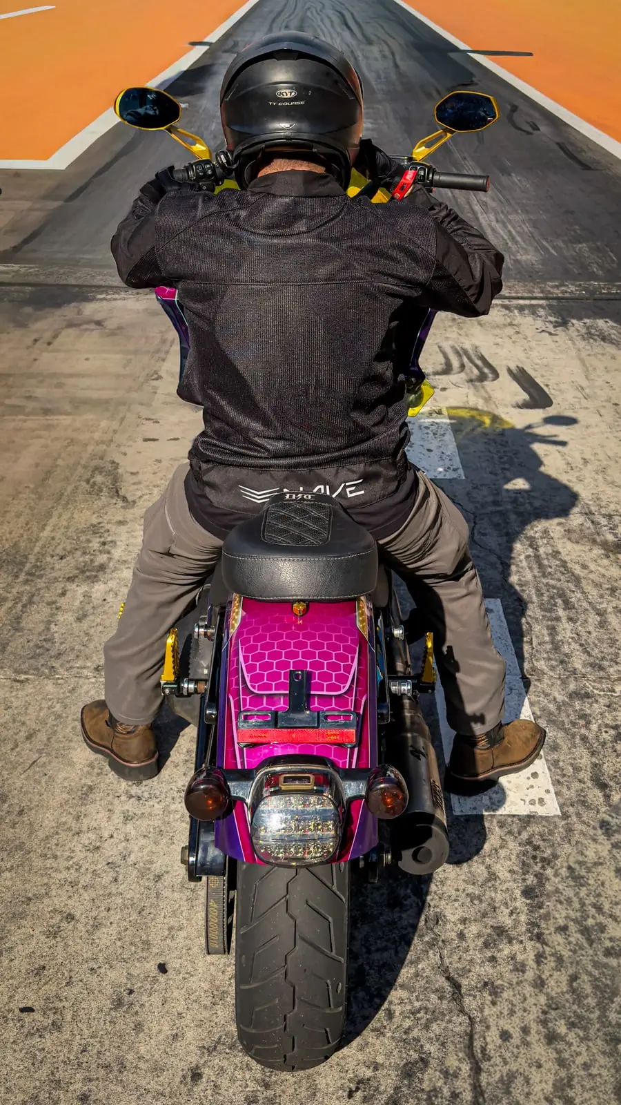 Piloto visto de cima acelerando uma moto customizada rosa e roxa pela pista de arrancada.