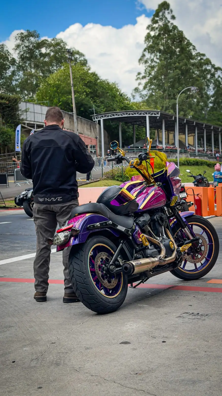 Moto customizada roxa e rosa parada na entrada da pista, com árvores e céu azul ao fundo.