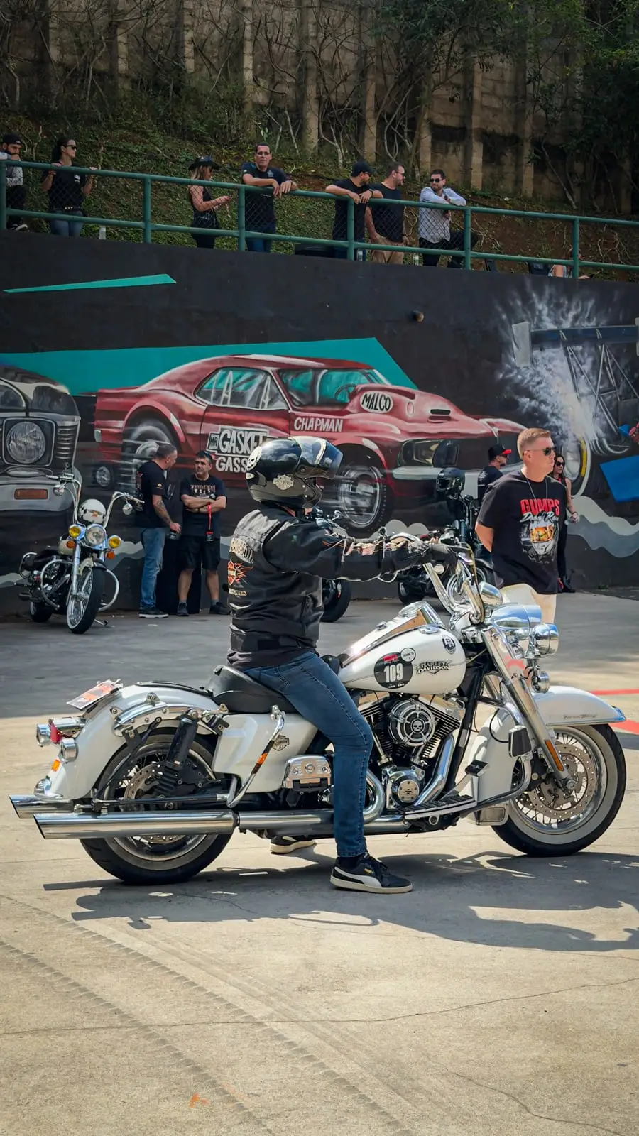 Piloto em uma Harley-Davidson branca em frente ao muro grafitado do autódromo.