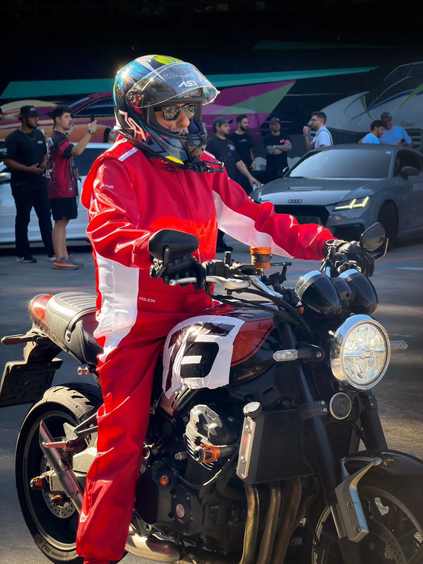Piloto de macacão vermelho e capacete parado sobre a moto, olhando para a pista antes da largada.