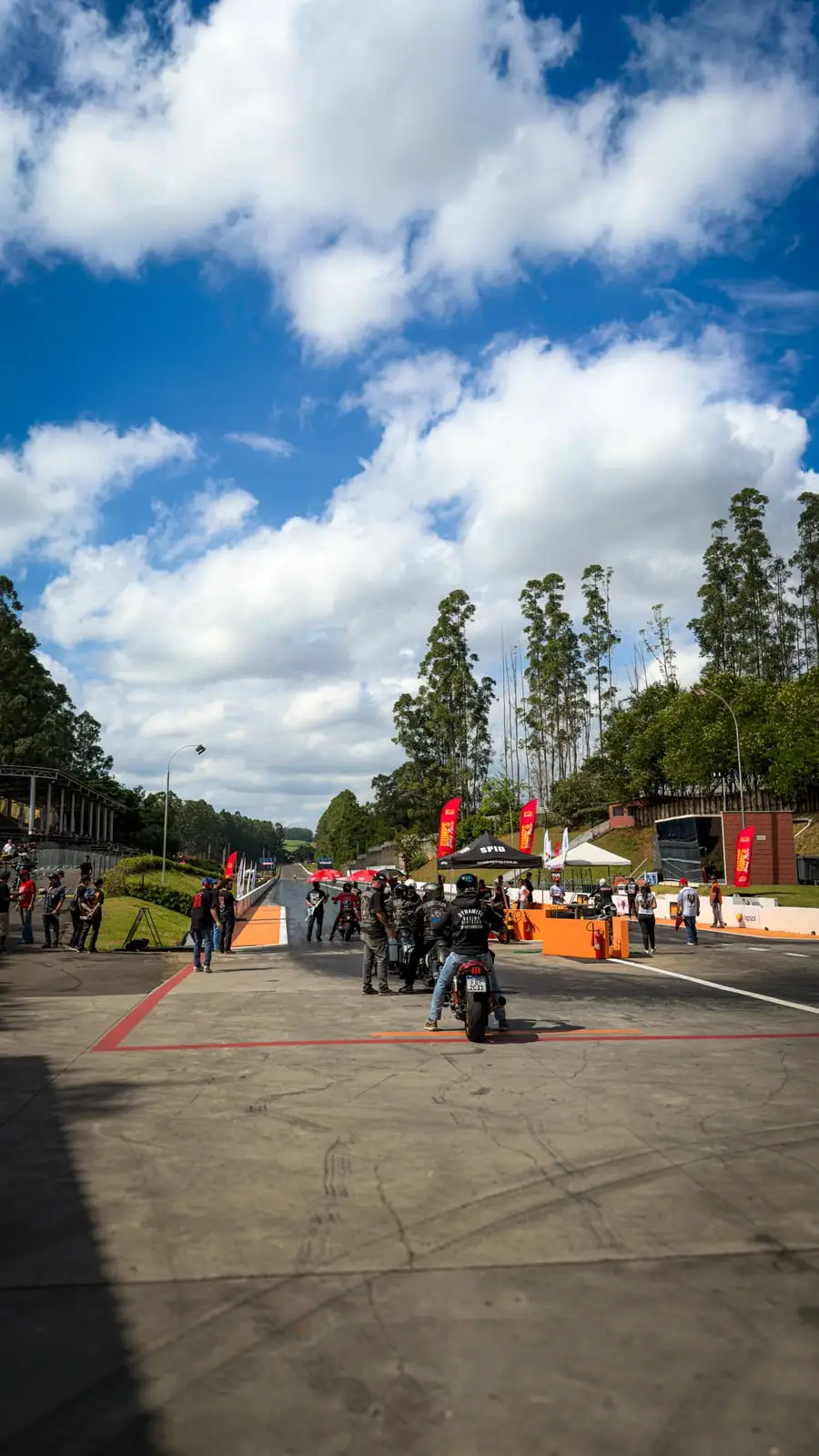 Vista aberta da pista de arrancada, com as equipes reunidas na largada e céu de nuvens altas.