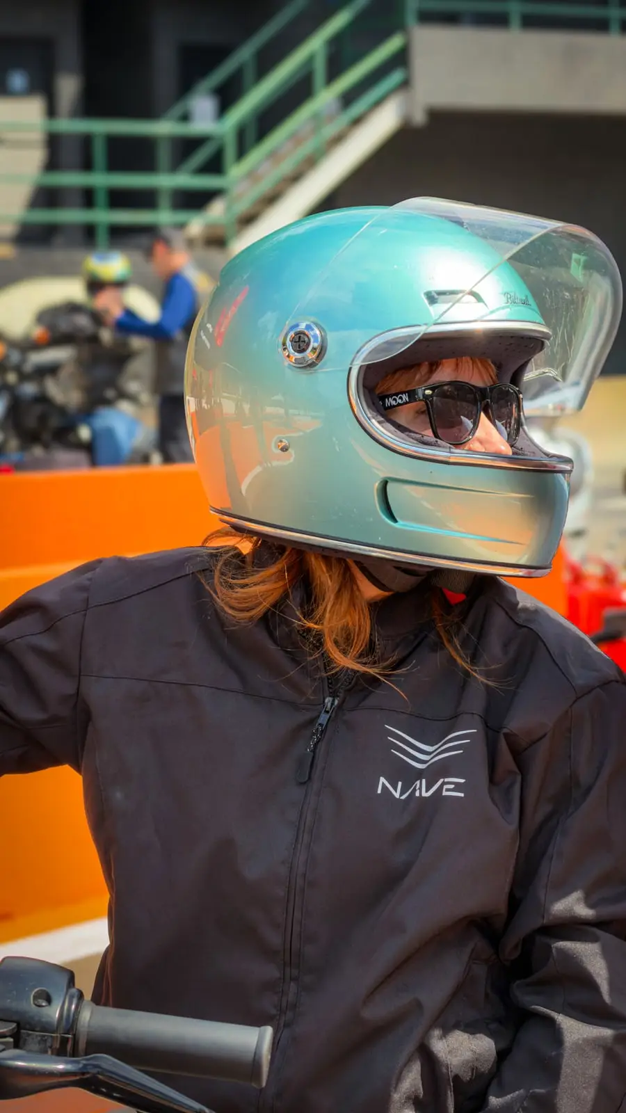 Retrato de pilota usando capacete verde metálico, com as motos e o público desfocados atrás.